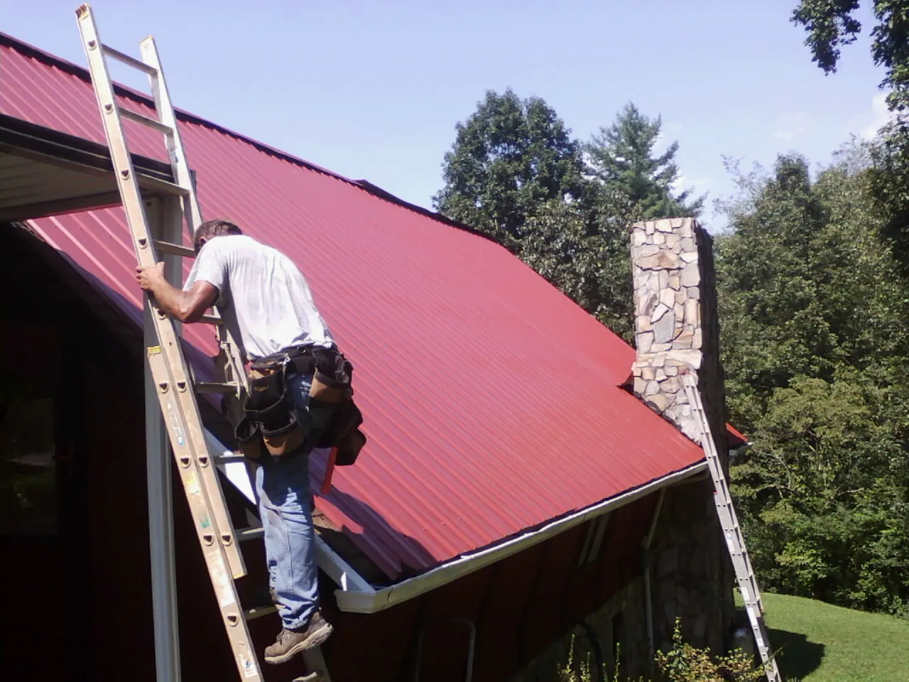 Roofer on a ladder beside a red metal roof and stone chimney — Soffit & Fascia Repair by Roofing Brothers in Mcloud